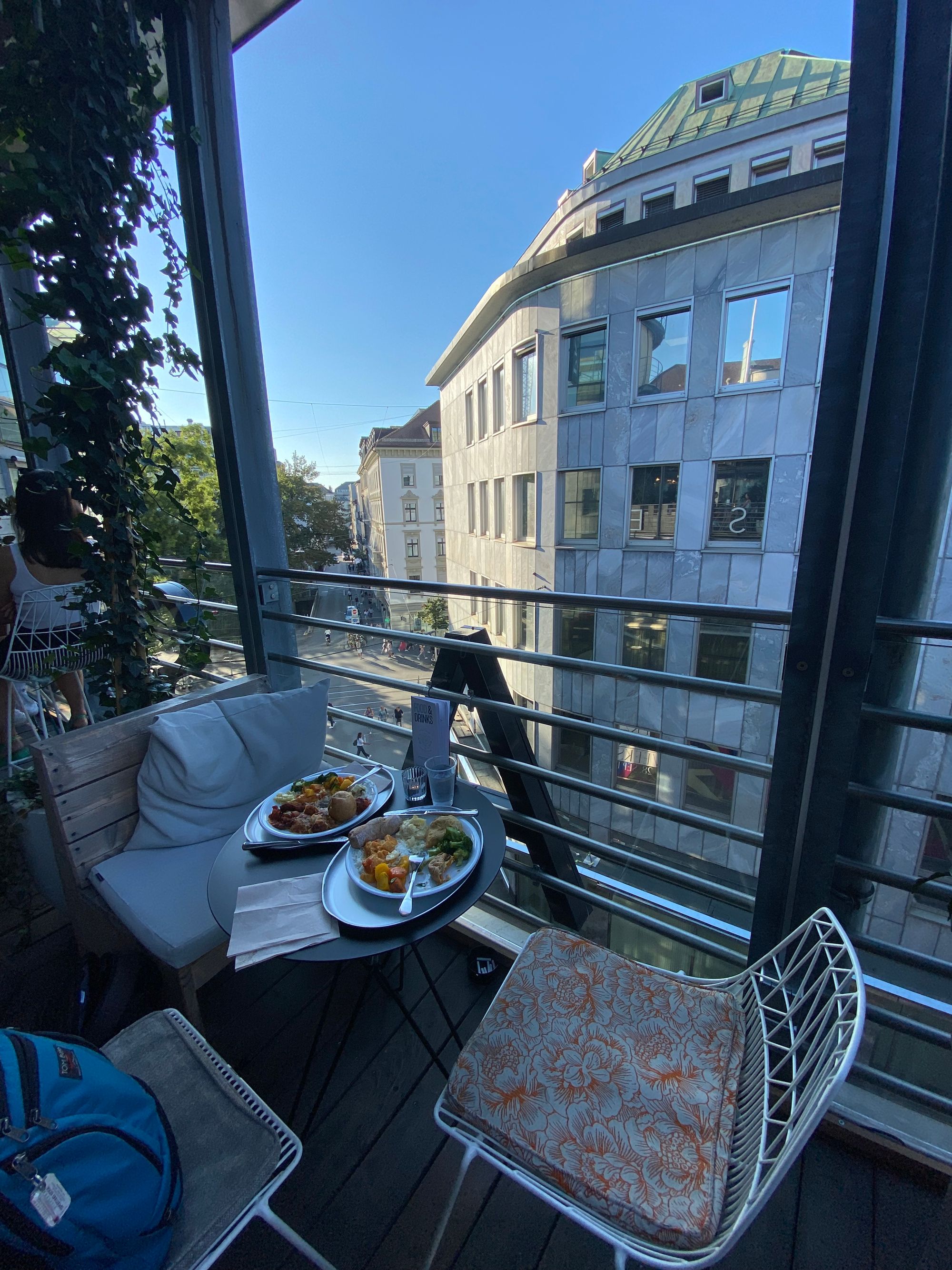 Two plates of food on a table with two empty chairs on a balcony overlooking a city street.