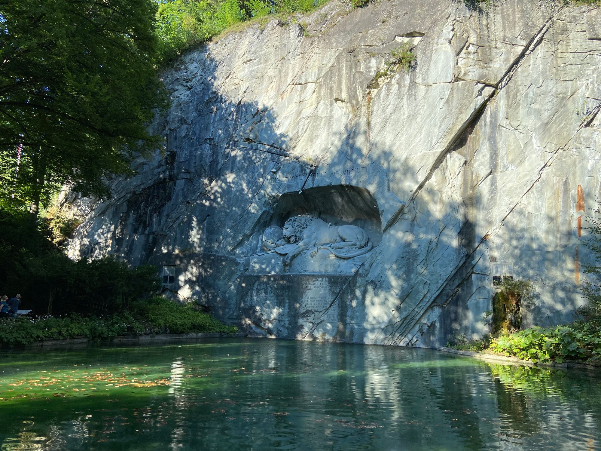 Sun dappled cliff face above a blue-green pond with a carving of a wounded lion in a niche about one-third of the way up the cliff.